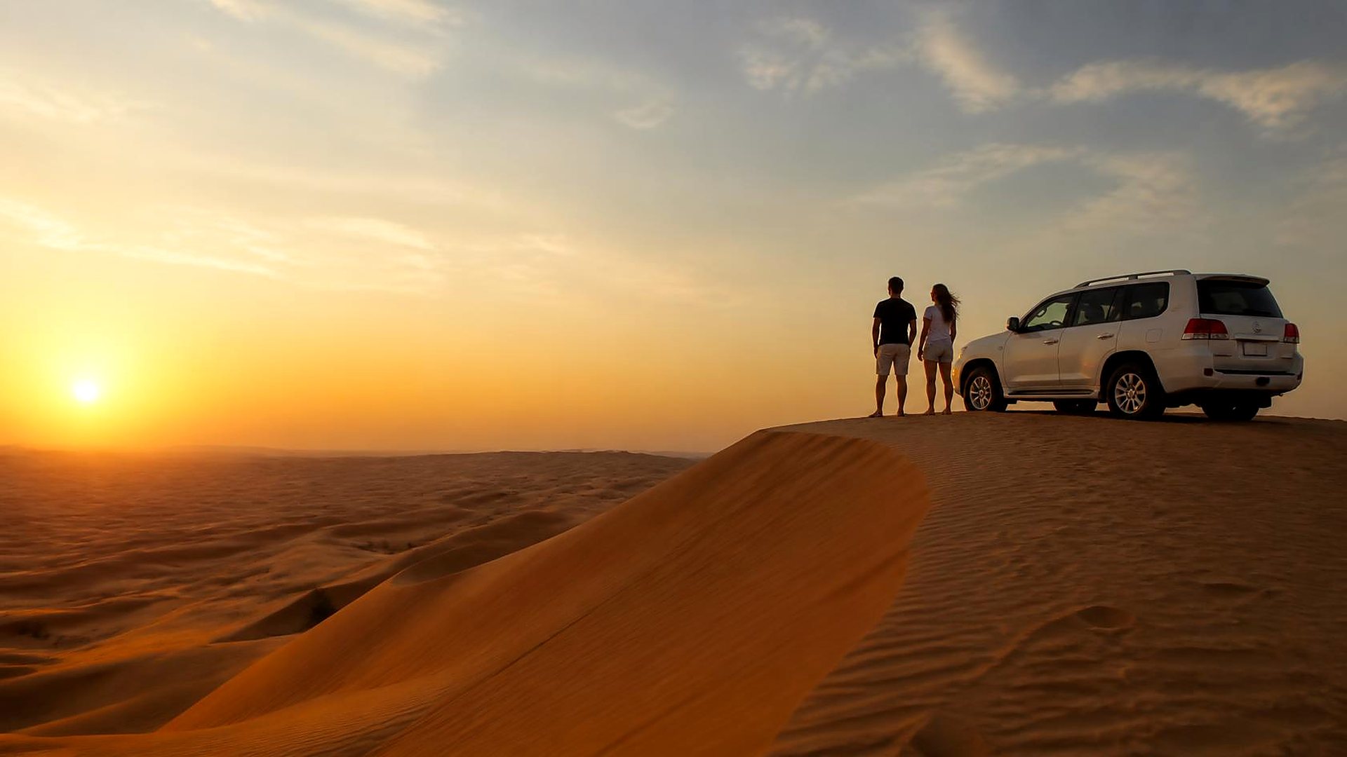 Couple standing on a desert dune beside a 4x4 at sunset