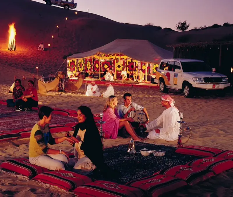 Guests on carpets by a campfire at a desert camp at dusk